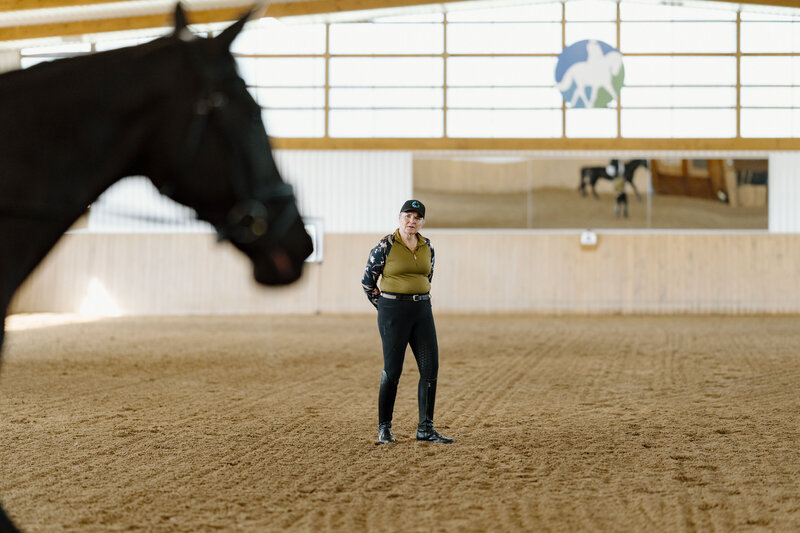Riding instructor watching her student ride by on a black horse in an indoor arena