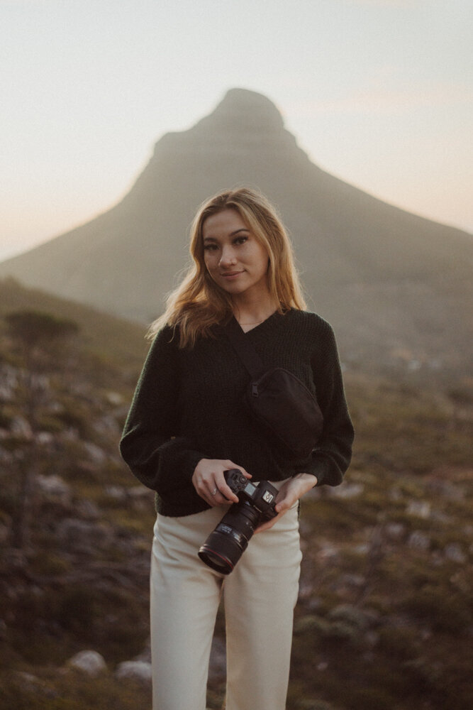 young woman holding a camera smiles with a mountain in the background