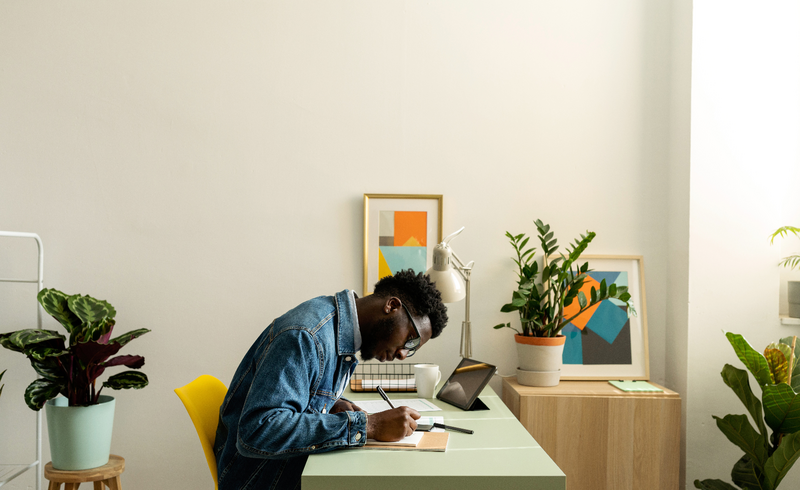 Young man with glasses and a denim jacket writing at a green desk, surrounded by vibrant houseplants and geometric artwork in a well-lit, modern home office.