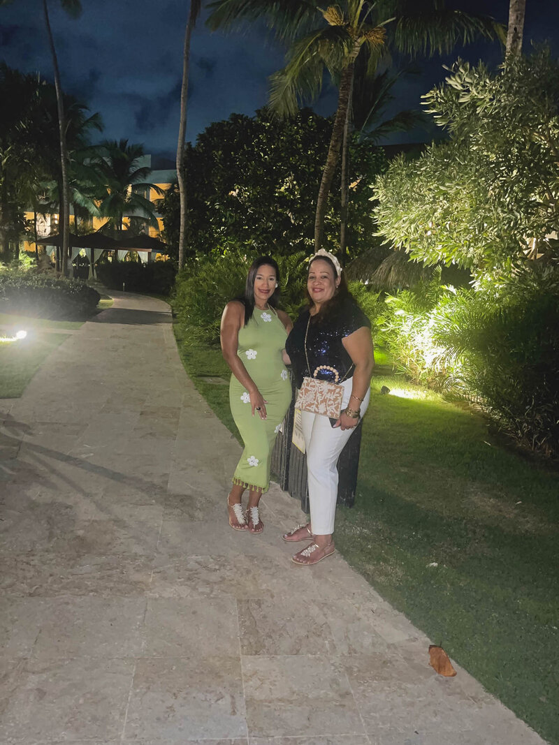 Two women smiling and posing together outdoors at night; one wearing a green dress with white flower designs and sandals, the other wearing a navy top with white pants and sandals, standing on a stone pathway surrounded by greenery.