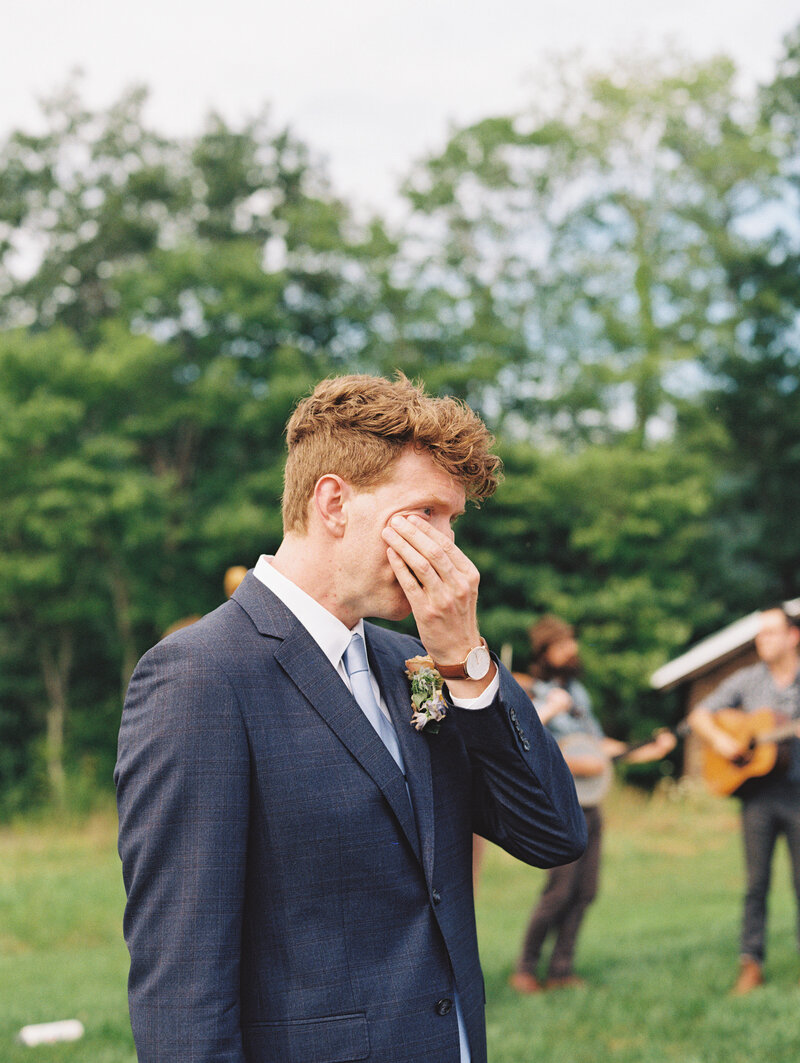 The grooms wipes tears from his face as his bride walks towards him at Paint Rock Farm in the hills of North Carolina, by destination photographer Megan Lynn of My Sun and Stars Co.