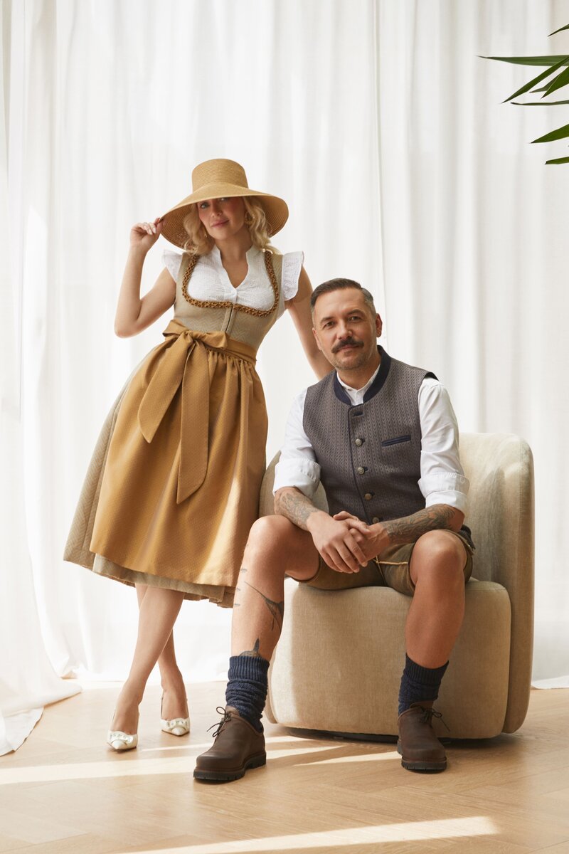 Stilvolles Trachtenpaar in moderner Designer-Tracht von Monaco Spatzl: Frau im gold-beigen Dirndl, Mann in Weste von Monaco Spatzl, Hirsch-Lederhose der Marke Schweigerts  – moderne bayerische Tracht aus München.