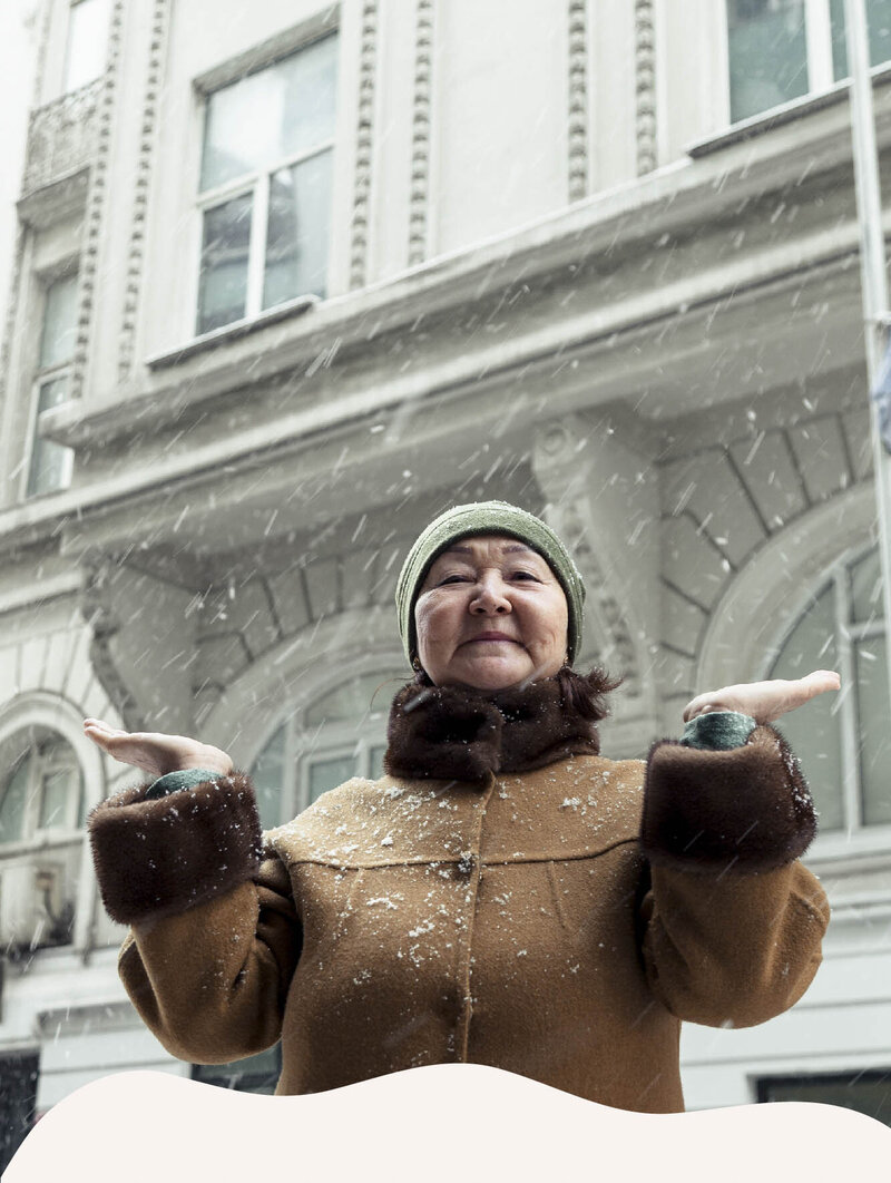 A feminine presenting person of color smiles gently down toward the camera, lifting their hands up to touch the falling snow. They are wearing a thick, brown coat and a green winter hat. The photo is framed by a snow-like design.