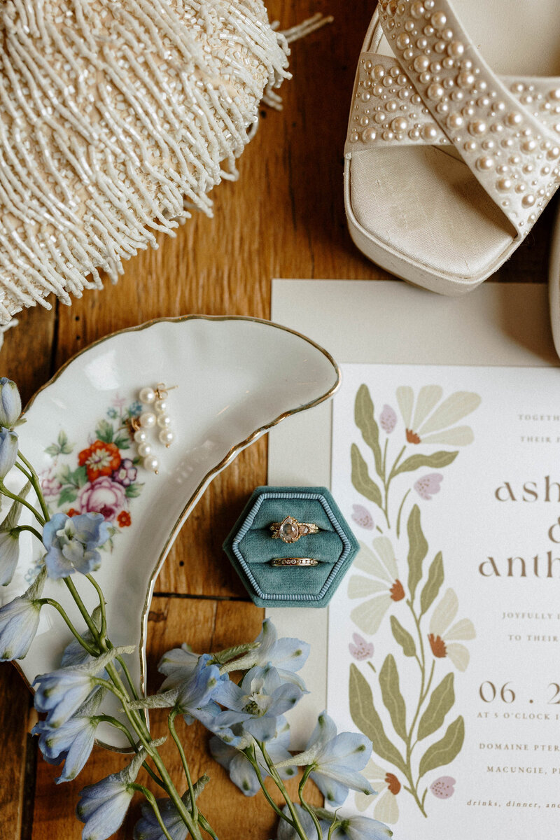 Elegant bridal accessories including pearl earrings, wedding rings in a teal box, floral plate, and wedding invitation on wooden surface.