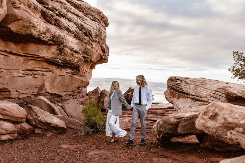 A couple walking through some red rocks during their sunrise elopement. The bride is wearing the grooms coat because it is chilly. They are holding hands and smiling. utah Elopement photographer