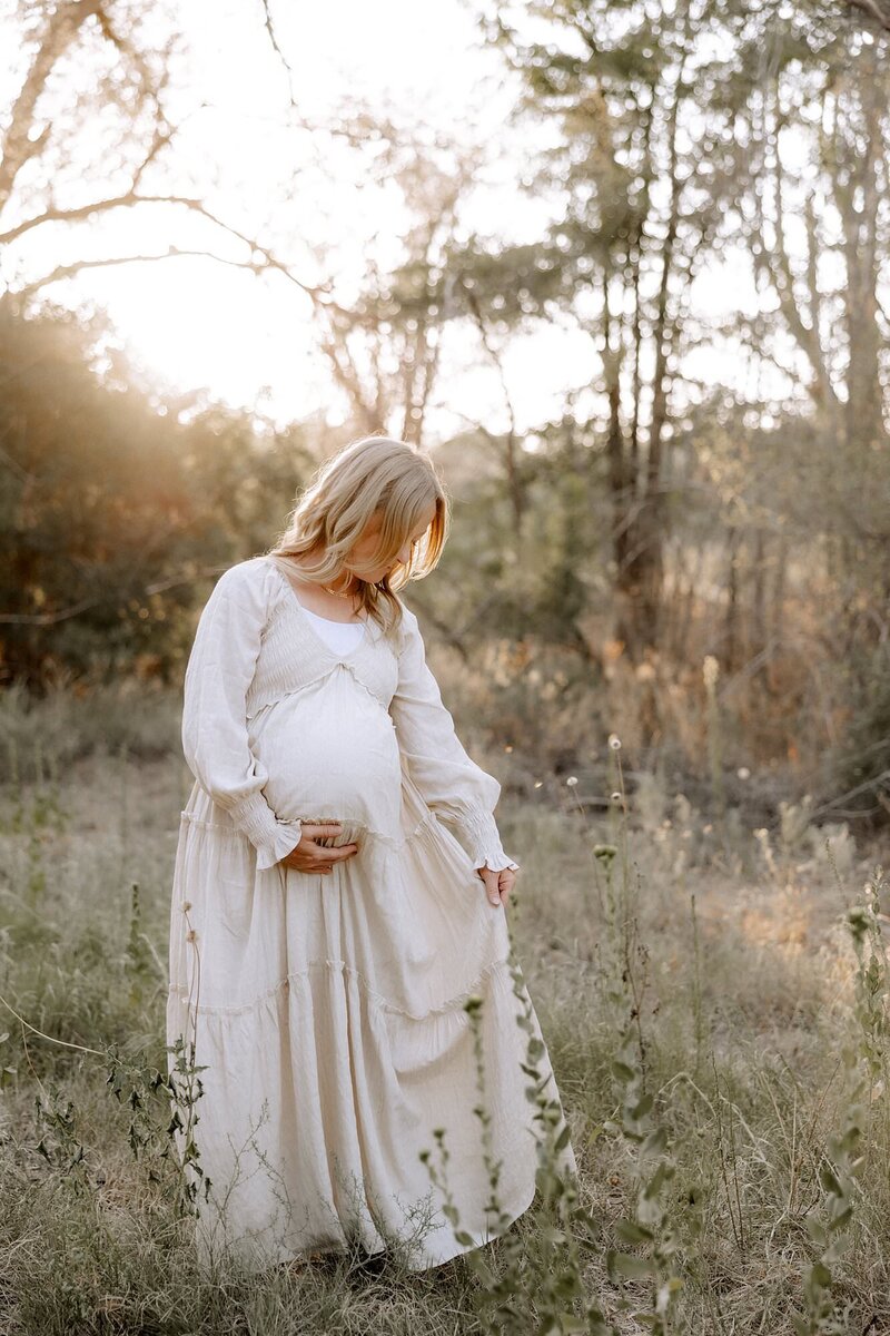 pregnant mama touching her lower belly area with one hand, swishing her dress with the other hand while looking down at her dress.