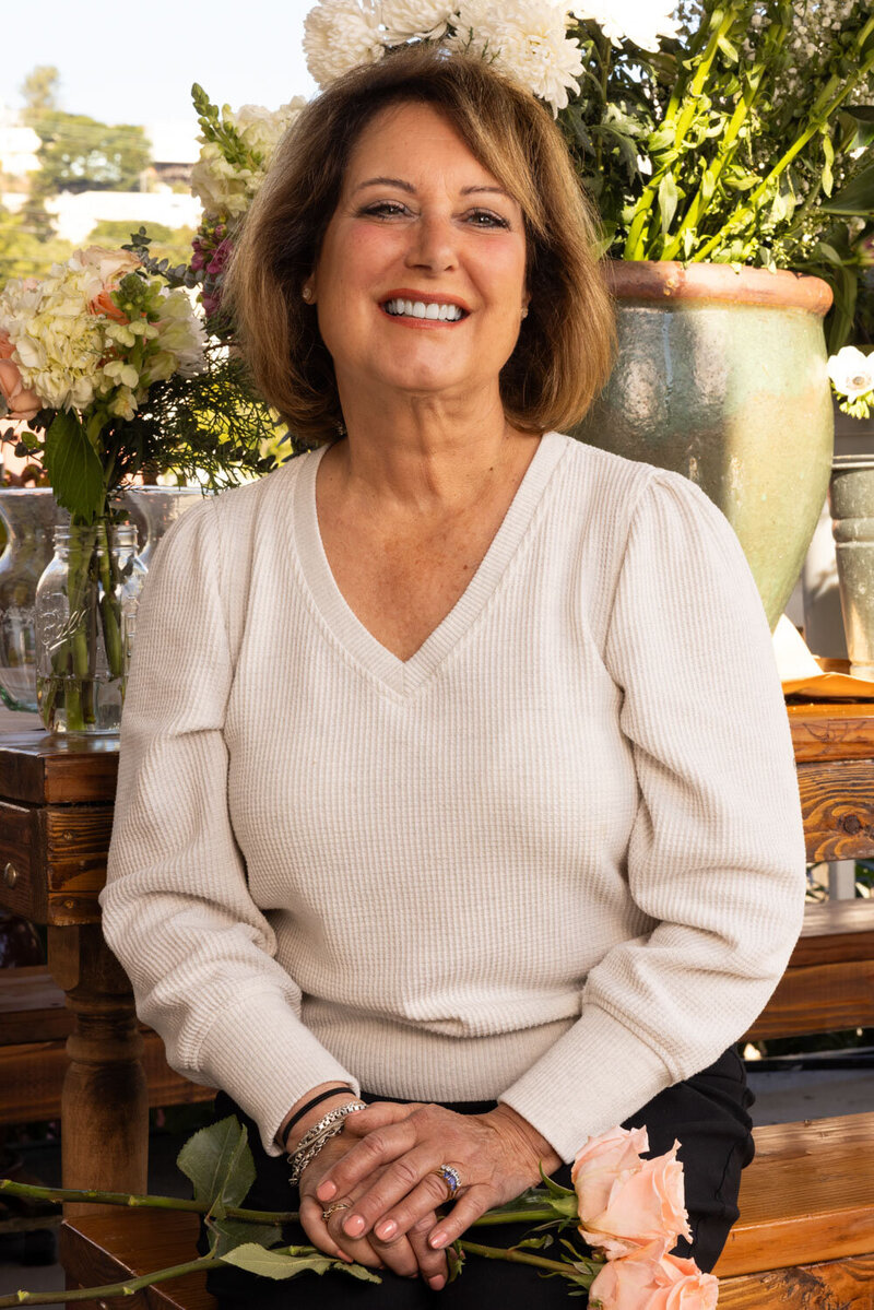 photo of Maria, the owner of Fleurish with Flowers, smiling while sitting among floral arrangements with a peach rose in hand