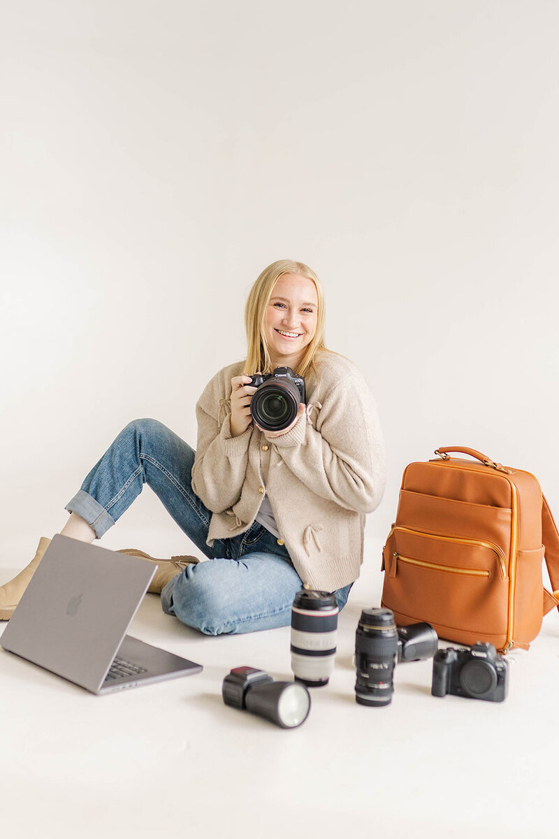 Jessica Nicole Photography smiling while holding a camera, surrounded by camera gear and a laptop, showcasing a passion for capturing beautiful moments in a stylish and inviting studio setting.