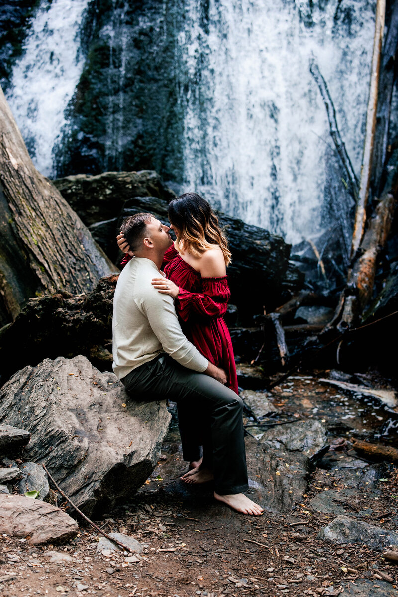 Man sitting on a rock with fiance standing in front of him embracing him in front of Kilgore Falls in Maryland