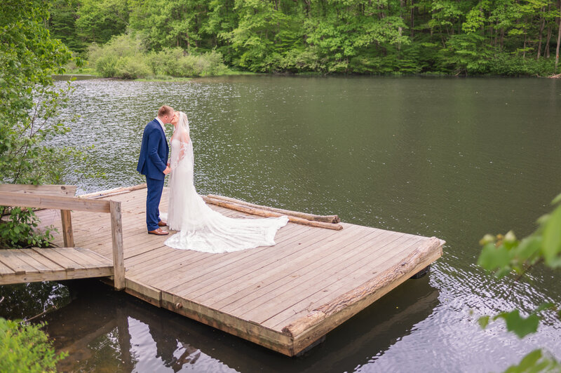 Bride and groom share a kiss after their first look on a wooden dock by a forest lake at a southwestern Pennsylvania wedding venue
