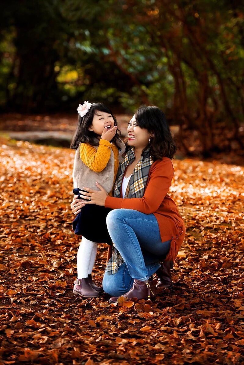 Mother and young daughter in yellow and orange laughing in the fall leaves at Queen Elizabeth Park.