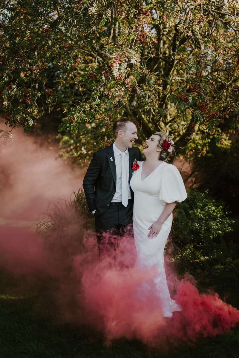 Bride and groom looking at each other laughing with red smoke around their feet