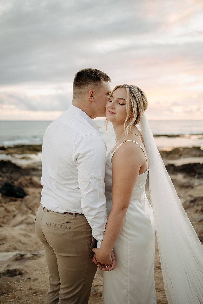 Groom kissing bride on the cheek while holding hands on the beach