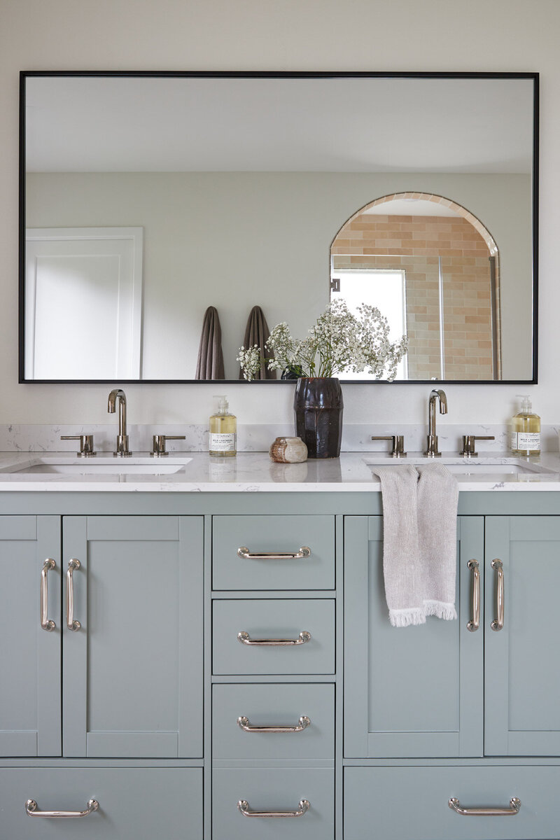 Bathroom with light blue bathroom vanity with silver cabinet pulls. A silver sink faucet with a black mirror above. Next to the sink is a dark brown vase with baby’s breath flowers, a Handsoap pump, and a white candle. Next to the vanity is a white wall with recessed white oak shelves with photos, jars, and a woven basket.
