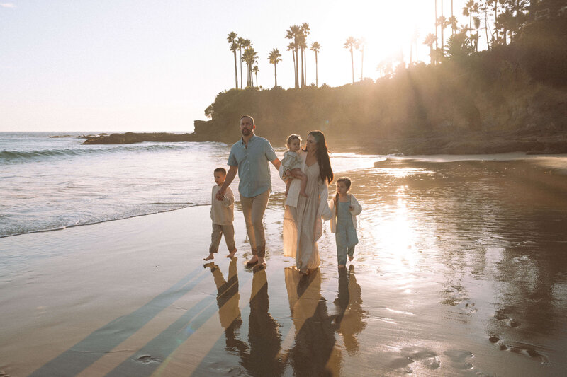 A family walking barefoot along the shoreline at Shaw’s Cove in Laguna Beach during golden hour, holding hands and smiling with the sun streaming through the palm trees behind them.