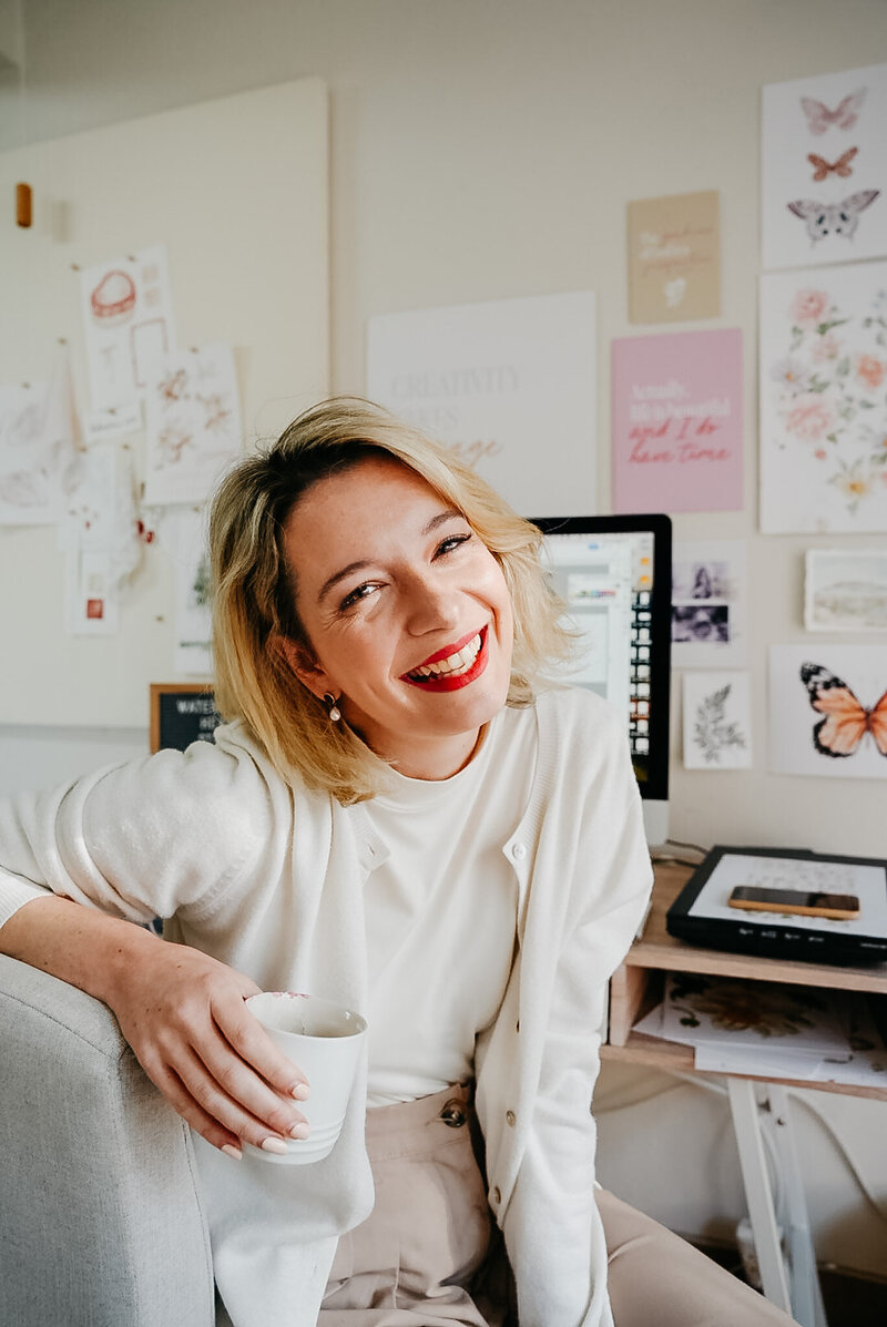 Jess Meyer smiling in her Watercolour Heart Illustration studio, holding a mug, with artwork on the wall and a computer and scanner behind her.