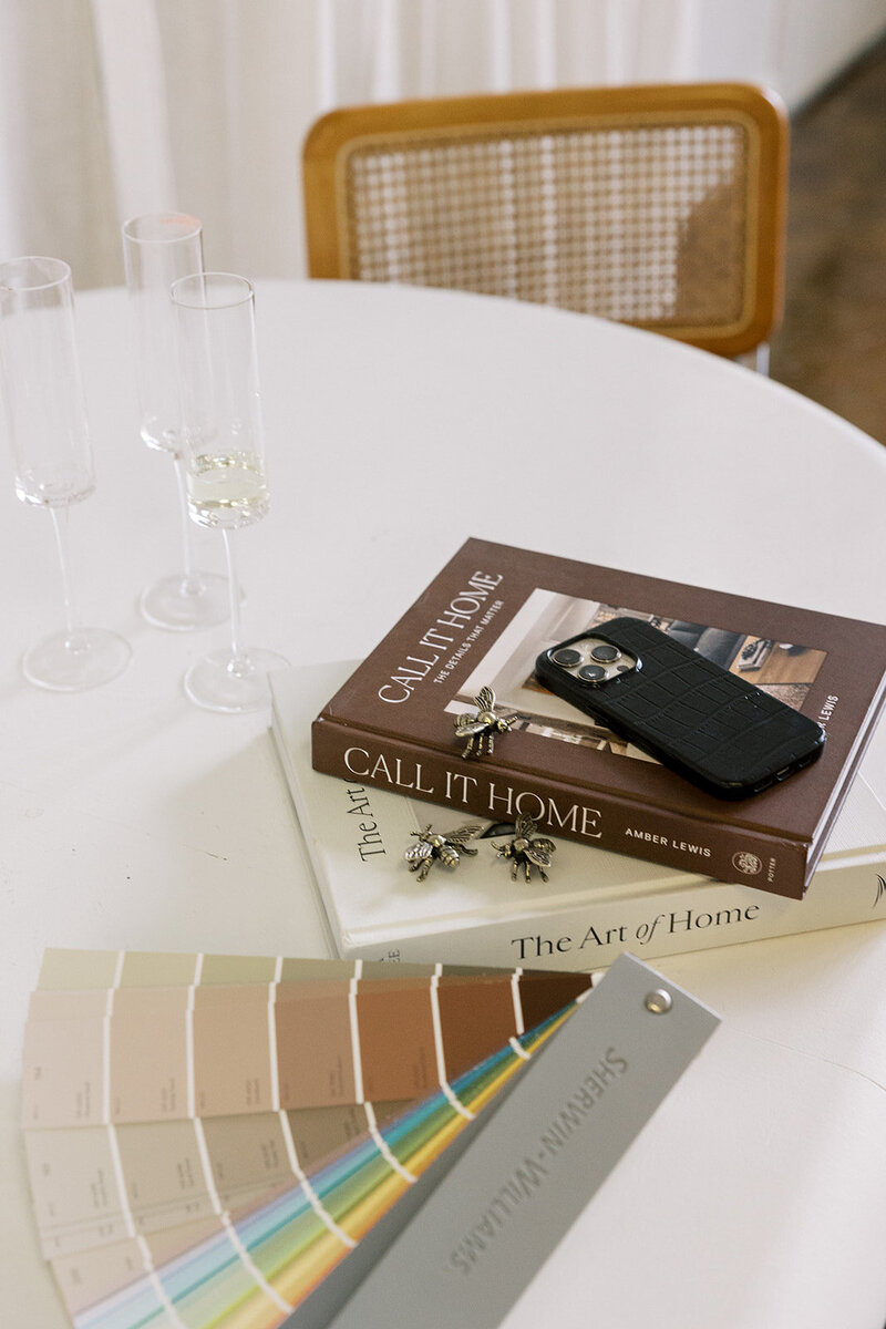 A round white table with champagne flutes, two stacked books titled "Call It Home" and "The Art of Home," a smartphone, decorative bee ornaments, and a Sherwin-Williams paint sample fan. A wooden chair is in the background.