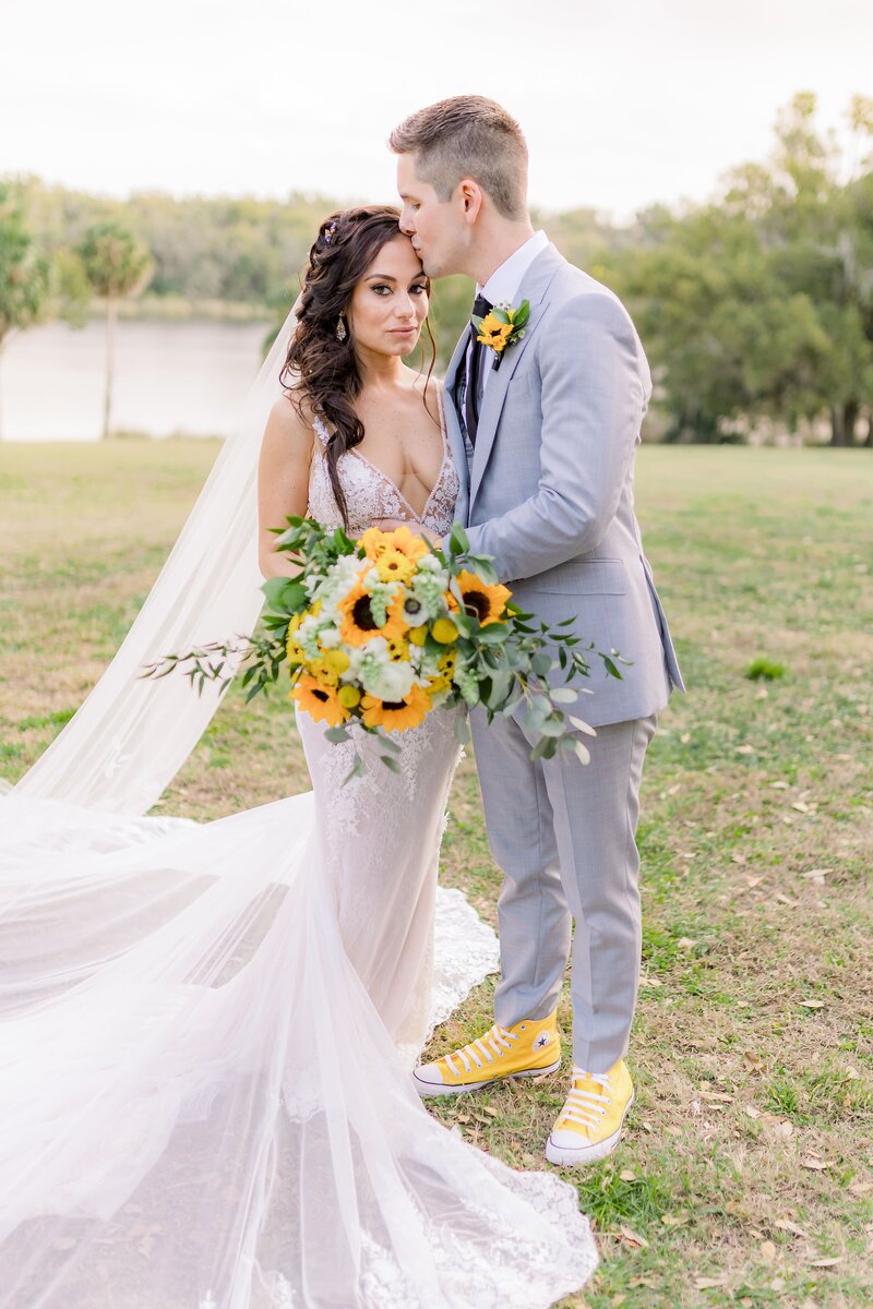 Bride looks at the camera holding sunflower bouquet as groom kisses her forehead