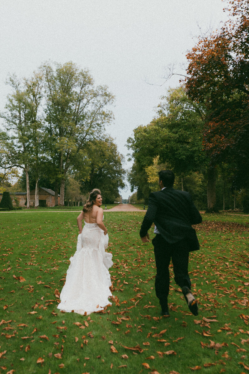 Bride and groom run across a green lawn scattered with autumn leaves, the bride looking over her shoulder in a candid moment.