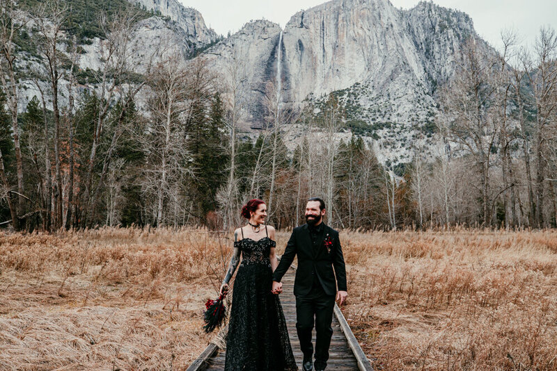A couple dressed in elegant black wedding attire walks hand-in-hand along a wooden pathway surrounded by tall grasses, with towering granite cliffs and Yosemite's majestic scenery in the background.