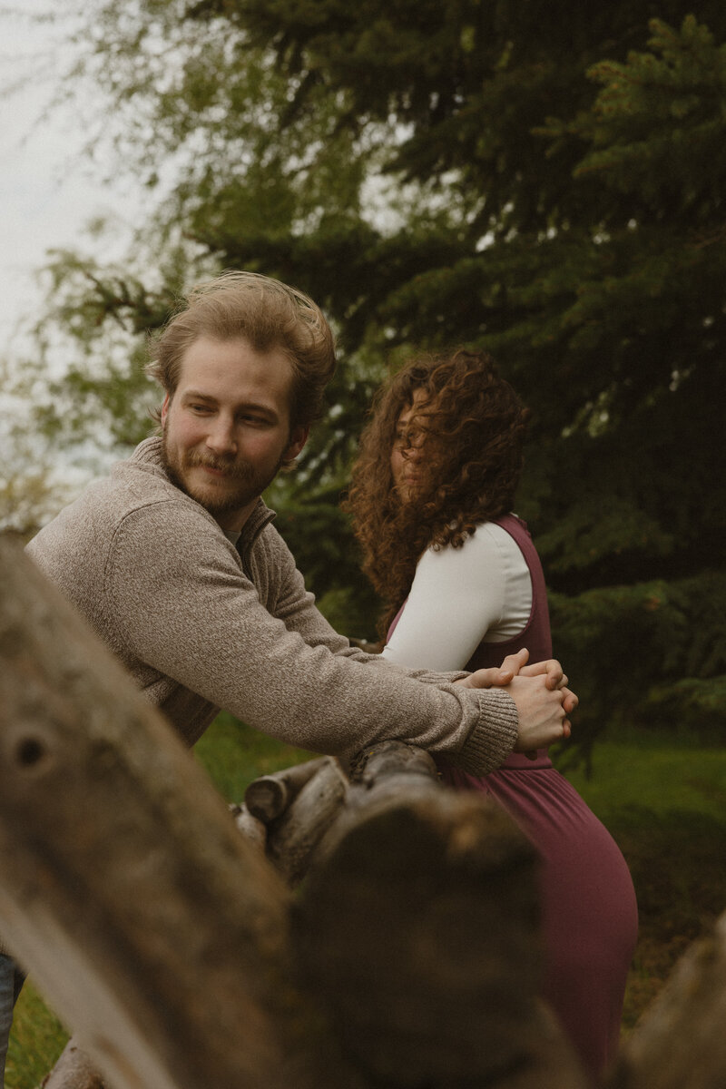 A girl sits on her fiance's lap in a grassy field during summer.
