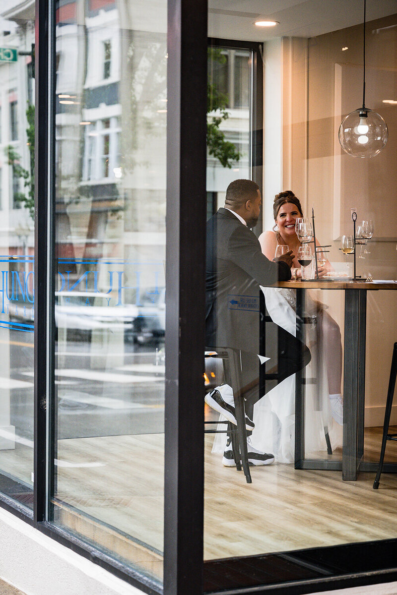 A newlywed couple on their elopement day sit inside of Wellhung Vineyard enjoying a wine tasting. The photo is taken from outside looking into the windows.