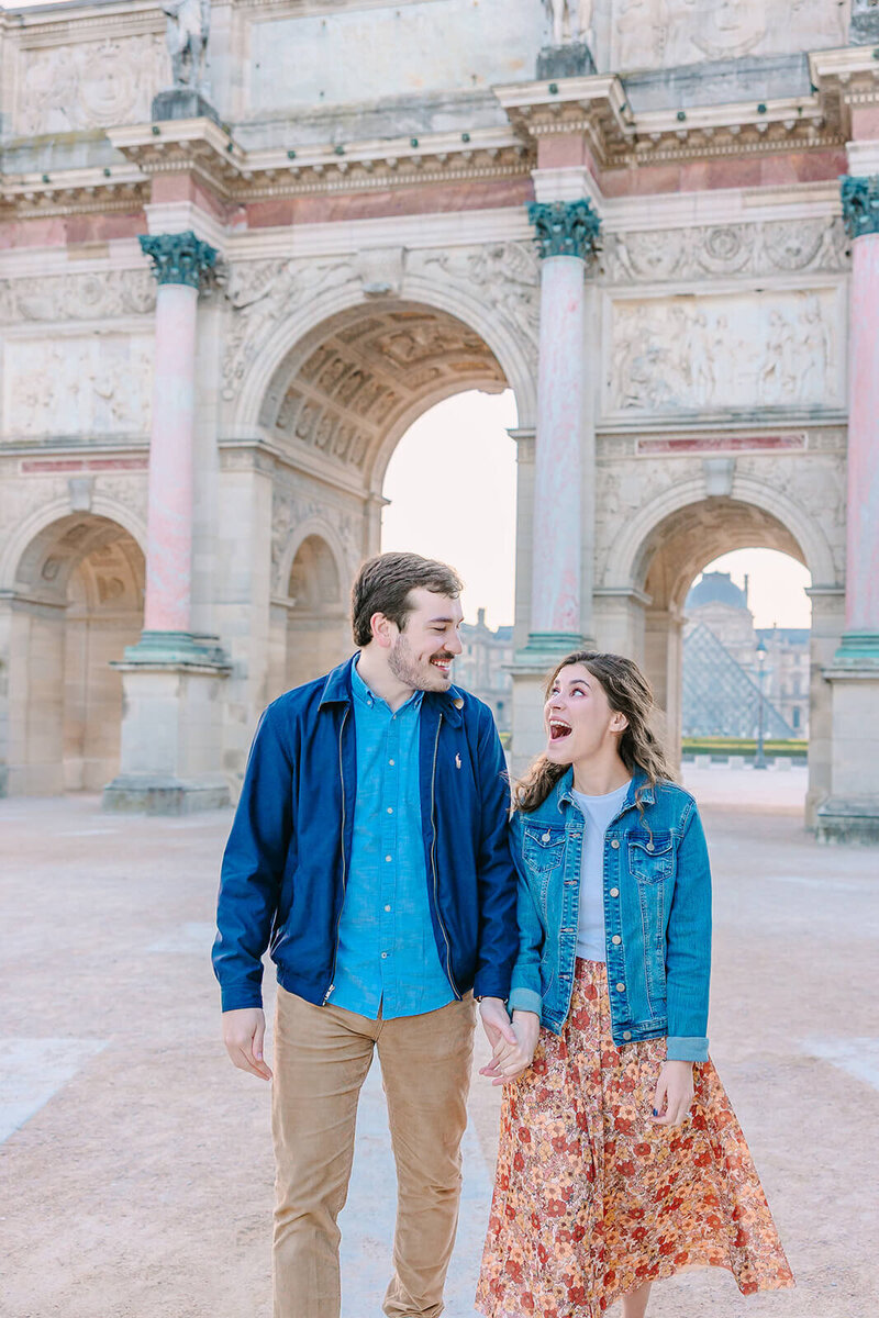 Man and woman holding hands during a Paris proposal at the Louvre and Tuileries Garden — the man smiling as the woman looks in excitement