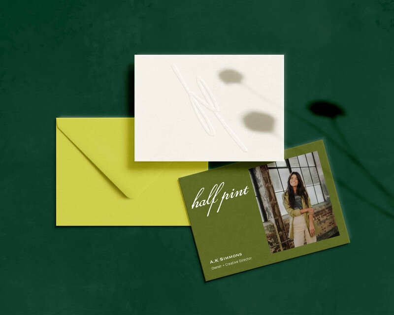 A business card for "half pint" featuring A.K. Simmons, crafted by a renowned branding designer, along with a blank white card and a lime green envelope, are displayed against a dark green background. Shadows of plant stems are visible in the background.