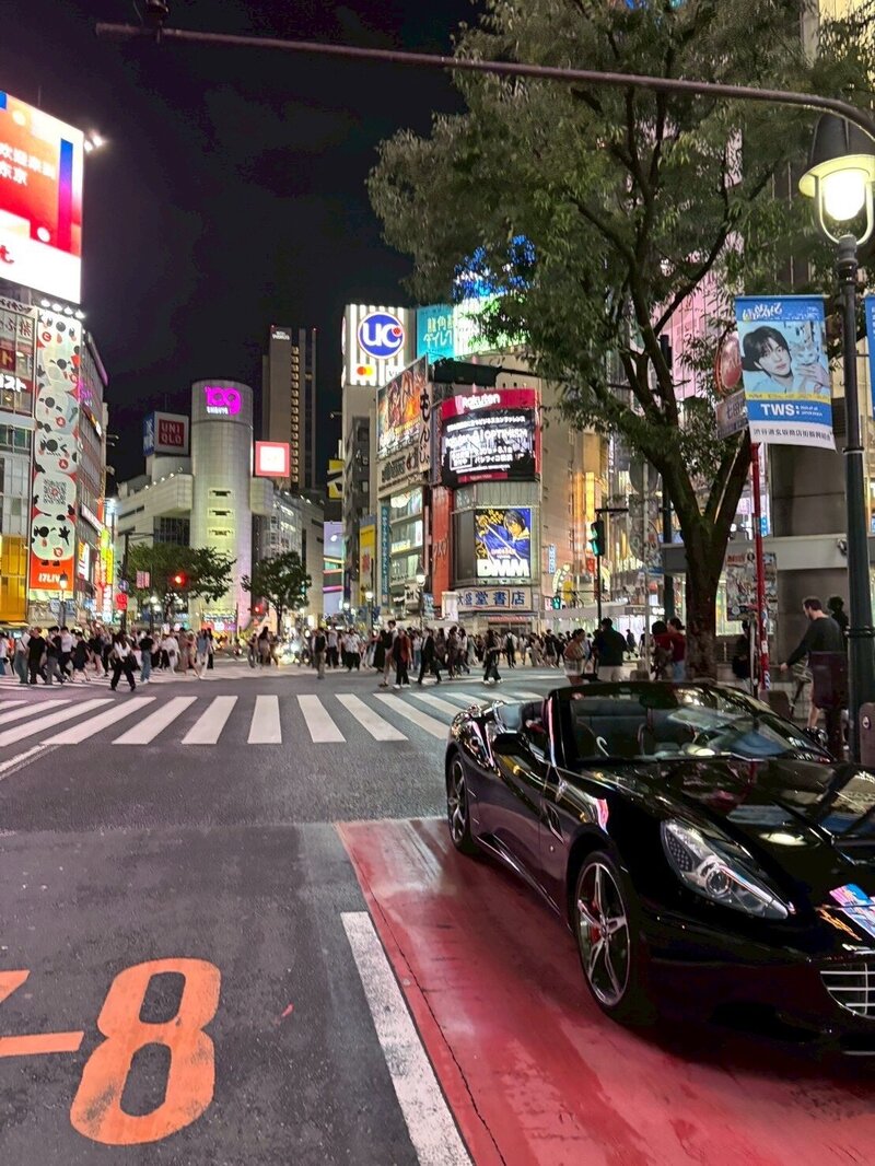A sleek black Ferrari convertible commands attention as it cruises through Tokyo’s iconic Shibuya Crossing, surrounded by towering billboards and bustling city vibes.