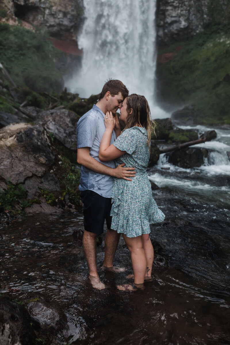 A red headed man wearing a blue button down shirt wraps his arms around his fiancée's waist, as they both stand forehead to forehead in the cold water just below Tumalo Falls in Bend Oregon for their adventure engagement session. | Erica Swantek Photography
