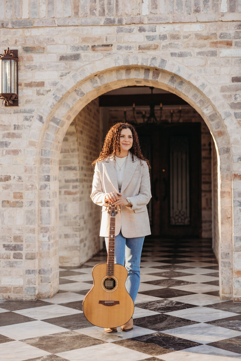 A young woman with curly, brown hair holding a guitar upright, standing between a brick arch on black and white marbled floor smiling off into the distance