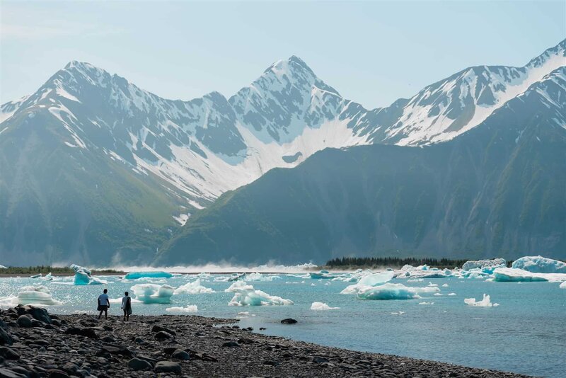 A couple, tiny in the framre, prepares to go for a polar plunge during day 2 of their luxury elopement package in Alaska.  In front of them is an expanse of a lagoon, filled with white and blue icebergs glowing in the sunlight.  Behind the lagoon, huge snow covered mountains fill the frame with bits of bright blue sky poking out between peaks. 
