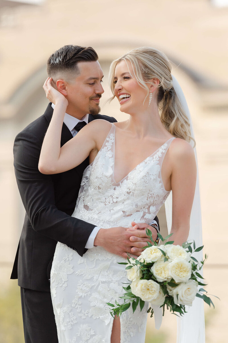 a bride in a lace dress cups her groom's face and laughs.  Taken outdoors at the NAC by Ottawa wedding photographer JEMMAN Photography