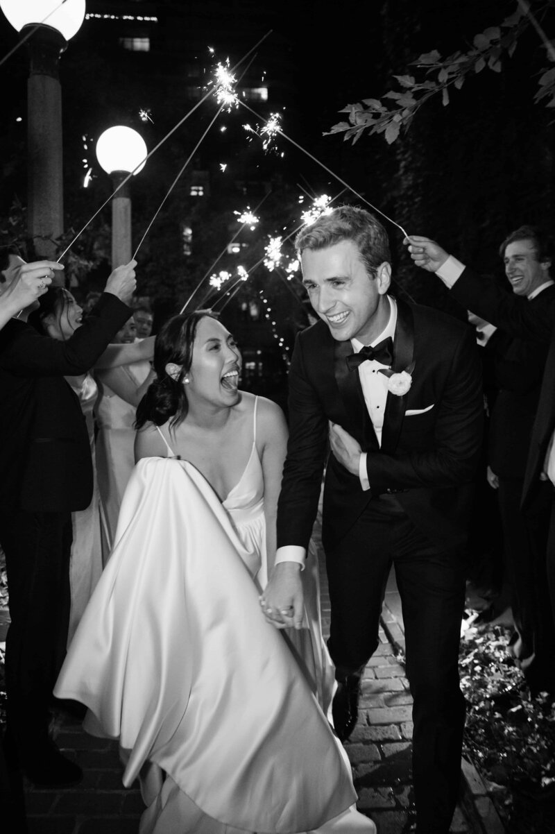Black and white wedding portrait of excited bride and groom surrounded by guest and sparklers