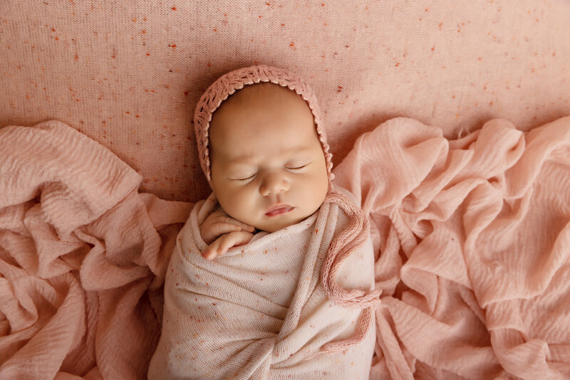Aerial image. Baby girl swaddled in peachy-pink for her brooklyn newborn photoshoot. Her hands are peeking out of the swaddle. She is wearing a matching bonnet and laying atop of matching layered and ruched fabrics. Captured by top Brooklyn, NY newborn photographer Chaya Bornstein Photography.