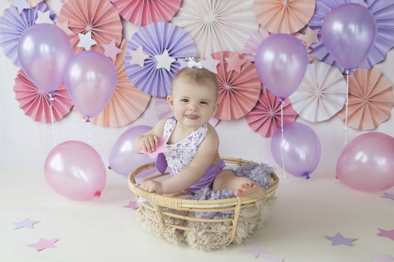 One year old girl sitting in a basket with purple balloons behind her