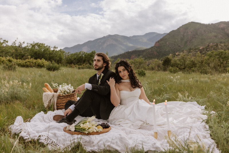Bride and groom sitting together on a picnic blanket in a mountain meadow, surrounded by wildflowers and candles, captured by Rainstorm Photo & Video in a romantic, cinematic elopement style.