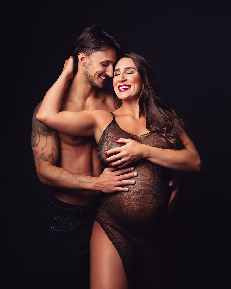 A brunette pregnant woman holding her shirtless husband and smiling together. Captured by Rosio Moyano, a greater Toronto maternity photographer.