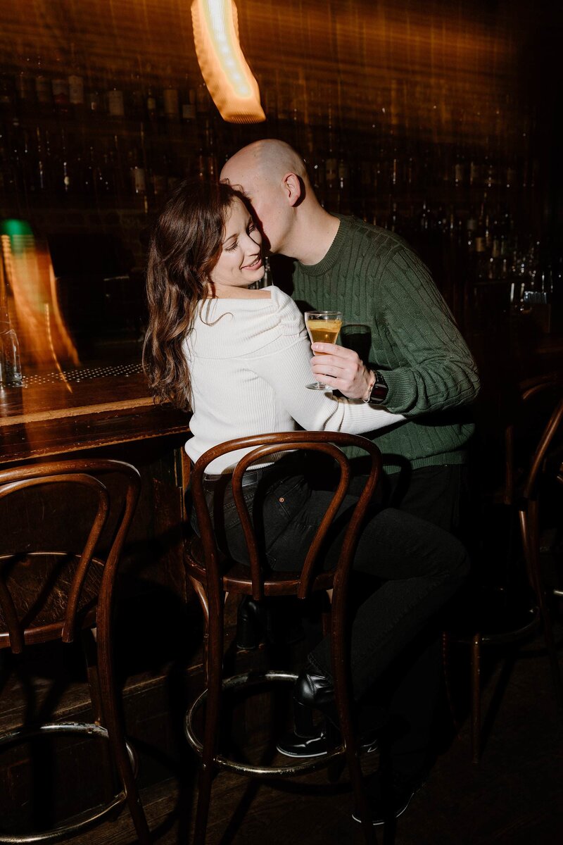 A couple sitting together at a bar as one kisses the other's cheek 