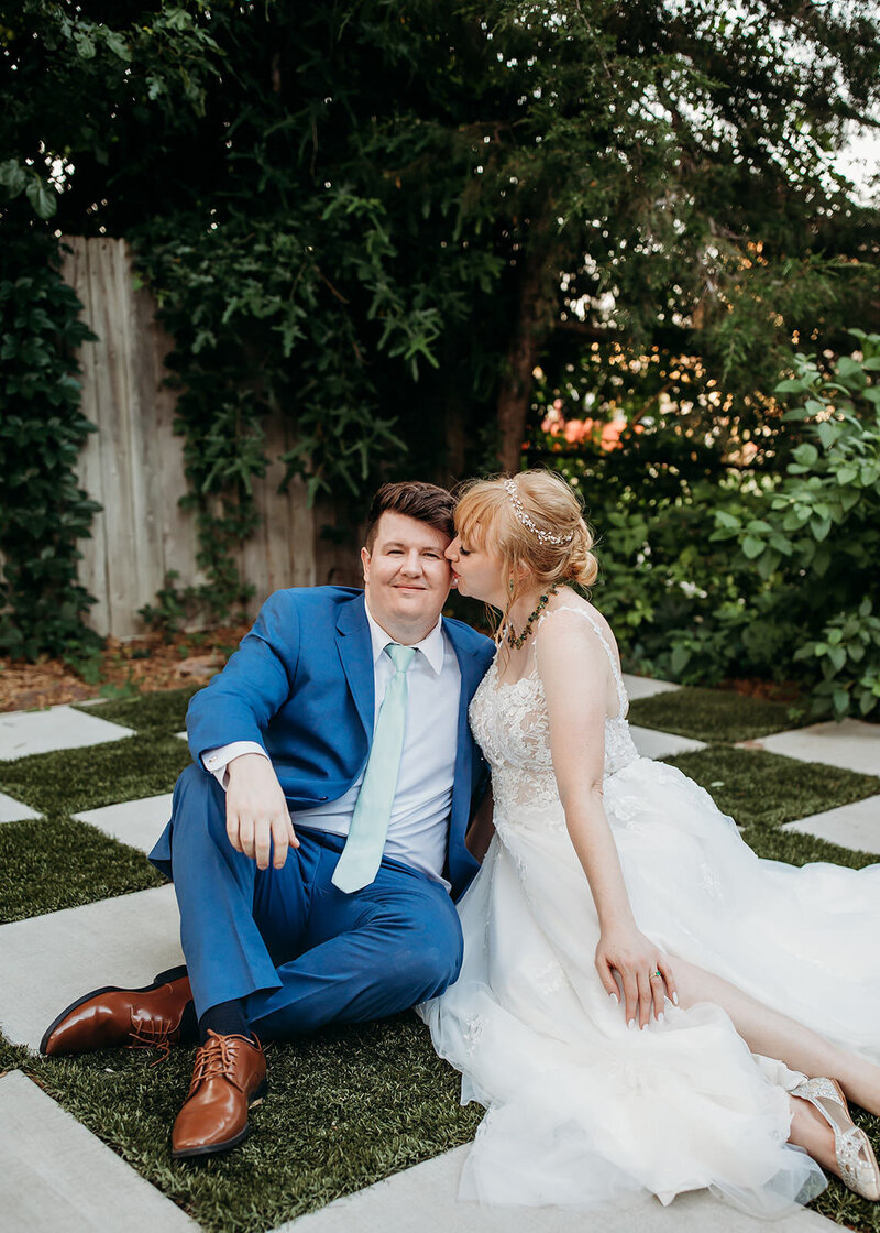 A newlywed couple shares a tender moment, with the bride kissing the smiling groom on the cheek, seated on patterned tiles amidst lush greenery