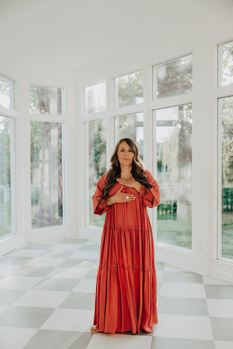 Patty in an orange dress in a room full of windows with her hands over her chest 