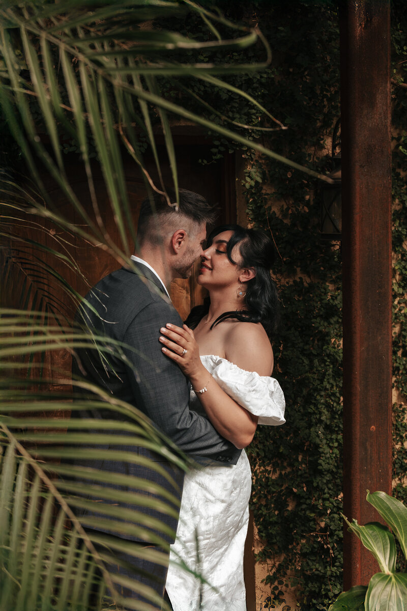 Couple sharing an intimate kiss in a romantic garden setting surrounded by palm fronds and wooden posts inside Villa Catalana’s greenhouse, perfect for elegant wedding photography.