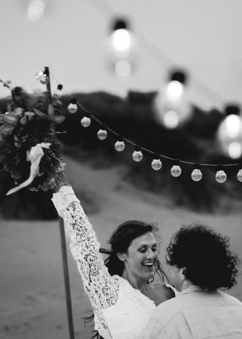 during their Oregon coast elopement, a couple in their wedding attire dances on the beach, under hanging  lights. they smile as the bride tosses her bouquet above her head as they dance.