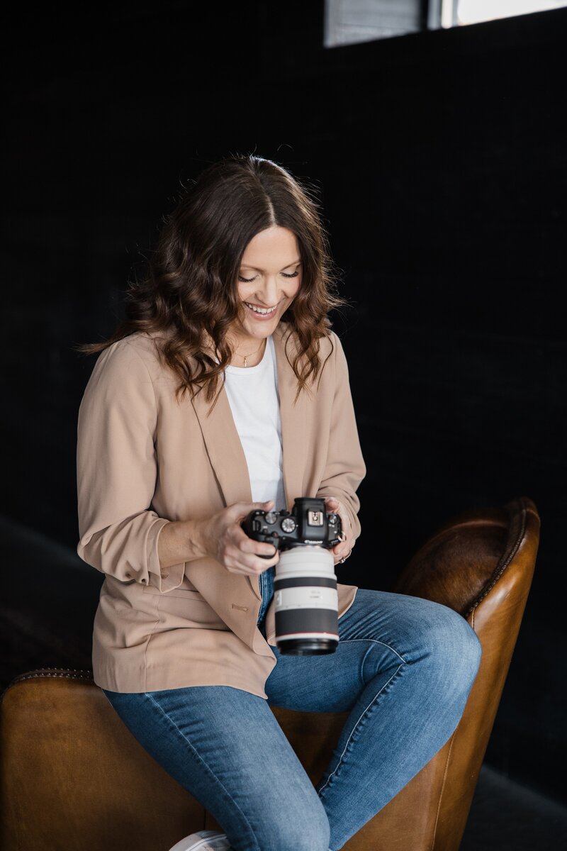 A person with wavy hair and a beige blazer sits on a brown chair, smiling and looking at the camera they are holding, embodying the essence of a talented photography website designer.