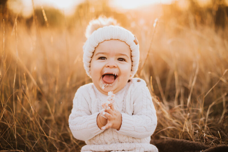 baby in a white crochet hat smiling and clapping with cake on her face
