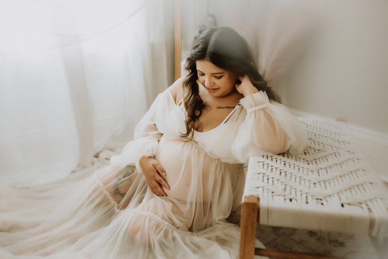 Pregnant woman in a sheer white dress sits on the floor, gently touching her belly, near a woven bench with soft lighting in the background.