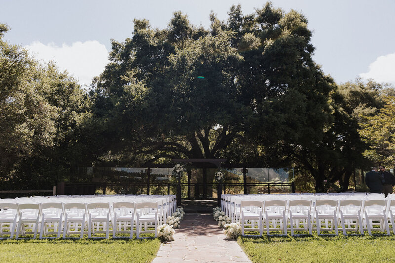 Nicole and Ivan’s outdoor ceremony setup with a large oak tree and white chairs, designed by Beyond the Event and photographed by Coyprint.