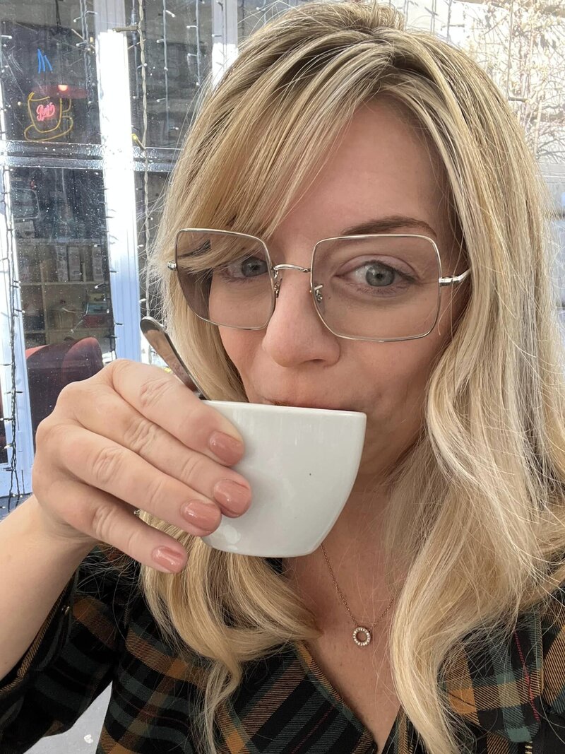 Marcie Muensterman has blonde hair and is wearing large glasses. She is sipping a drink out of a white mug.