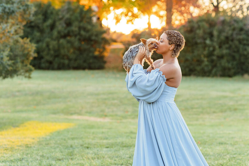A pregnant woman in a blue dress kissing her dog at sunset at Sully Historic Site.