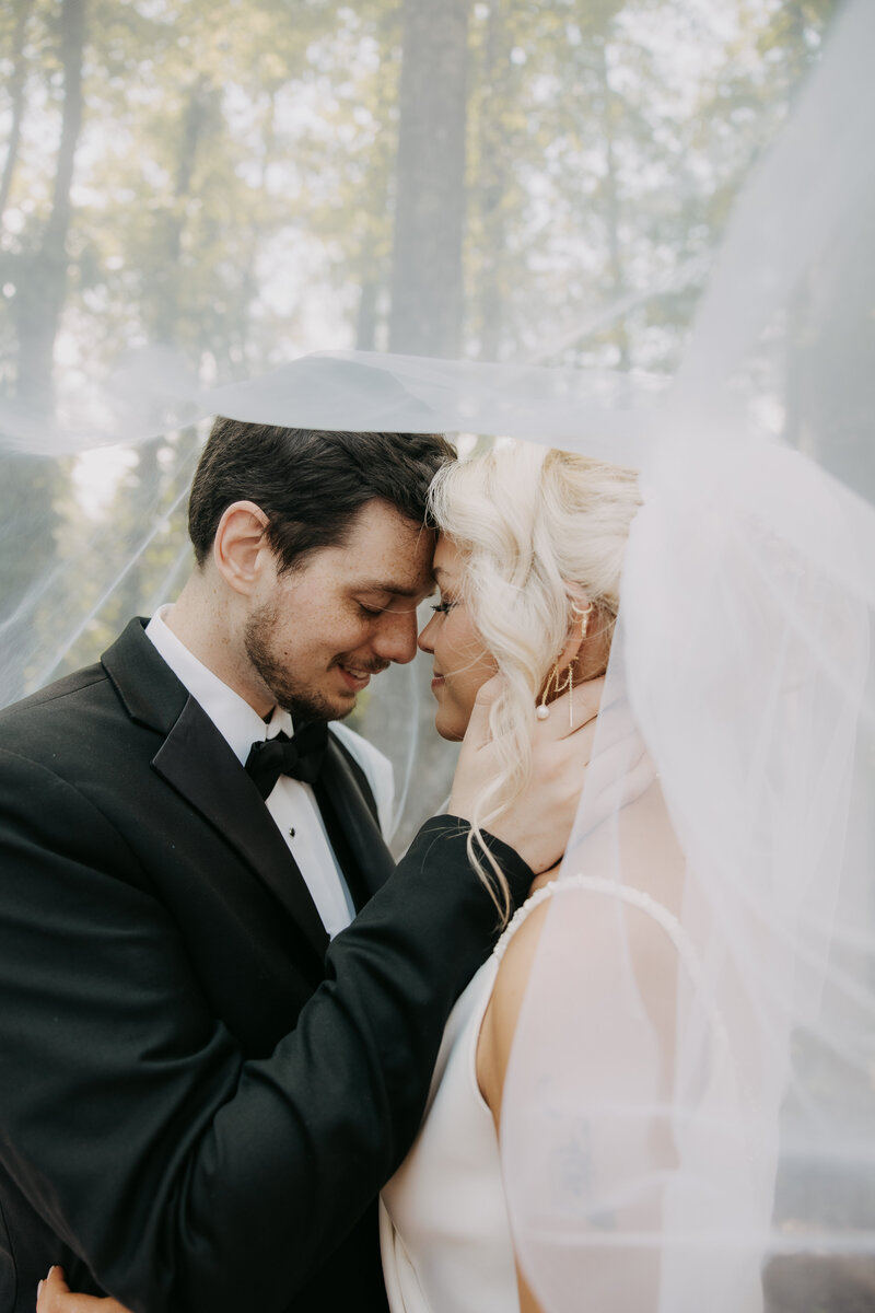 Groom holding bride's face, both eyes closed, with her veil gently flowing in front of the camera, standing by greenery at Kincaid Manor.